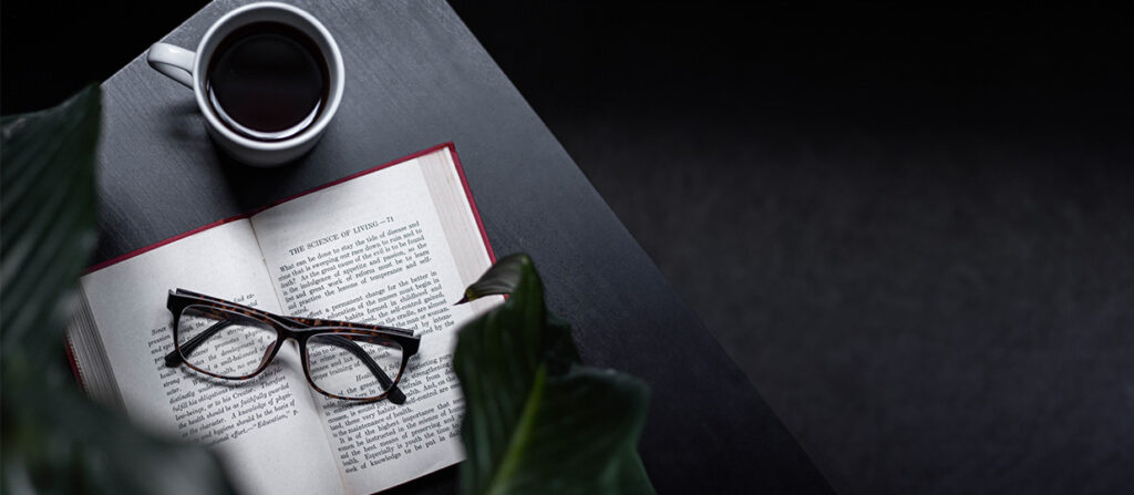 Picture of a flatlay of coffee, a book with reading glasses on top on top of a black table with a black background